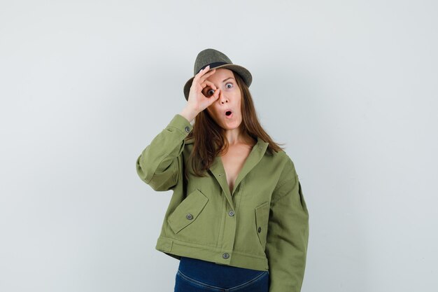 Giovane donna che mostra segno giusto sull'occhio in giacca pantaloni cappello e guardando stupito