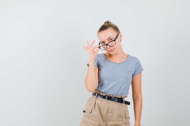 Giovane donna che osserva sopra gli occhiali in t-shirt, pantaloni e sembra sensibile, vista frontale.