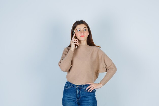 Giovane donna che parla al cellulare in maglione beige, jeans e sembra pacifica. vista frontale.