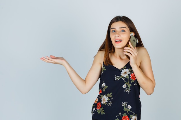 Giovane donna che parla al telefono cellulare, allargando il palmo della mano in camicetta e guardando perplesso, vista frontale.