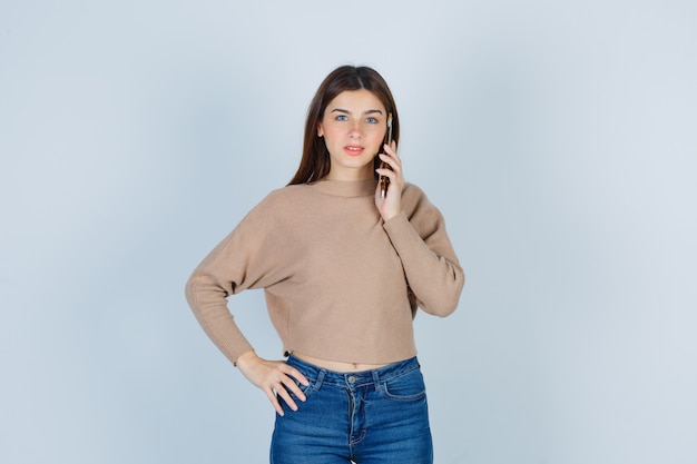 Giovane donna che parla al telefono cellulare in maglione beige, jeans e guardando allegro, vista frontale.