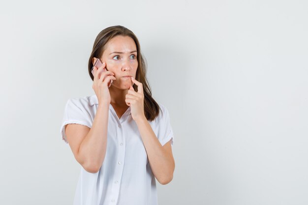 Giovane donna che parla al telefono in camicetta bianca e sembra persa