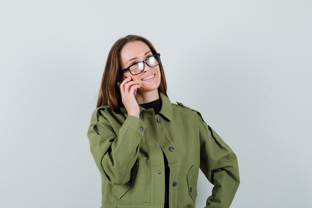 Giovane donna che parla al telefono in giacca verde, occhiali e che sembra felice, vista frontale.