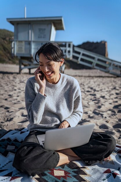 Giovane donna che parla su smartphone mentre utilizza il suo laptop in spiaggia