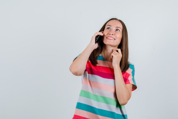 Giovane donna che parla sul telefono cellulare in maglietta e che sembra felice, vista frontale.