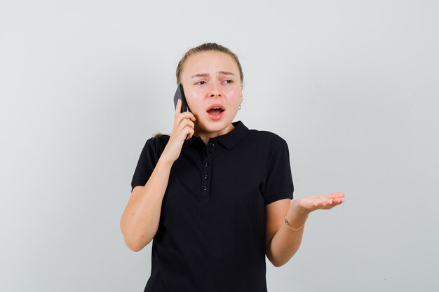 Giovane donna che parla sul telefono e alzando una mano in maglietta nera e guardando frustrata