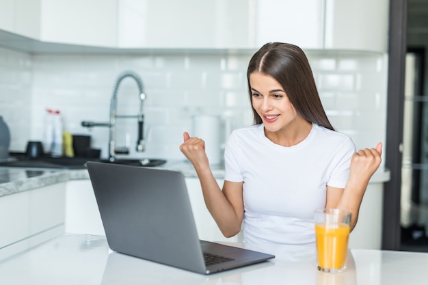 Giovane donna che per mezzo del computer portatile del computer alla cucina che grida fiero e che celebra vittoria e successo molto eccitati