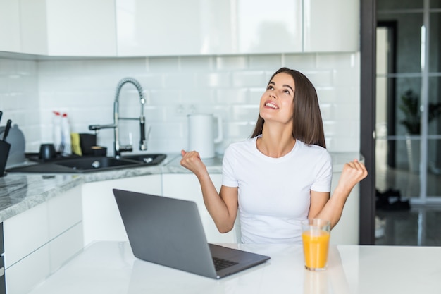 Giovane donna che per mezzo del computer portatile del computer alla cucina che grida fiero e che celebra vittoria e successo molto eccitati