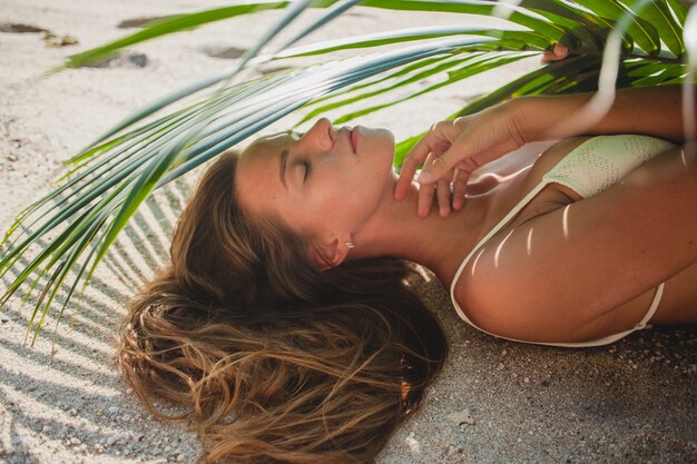 Giovane donna che pone sulla spiaggia di sabbia sotto la foglia di palma