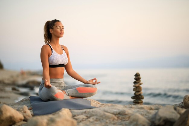 Giovane donna che pratica esercizi di rilassamento Yoga e meditando su una roccia in spiaggia. Copia spazio.