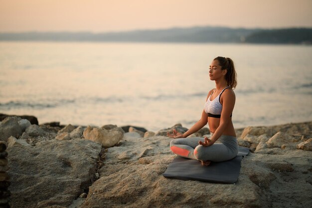 Giovane donna che pratica Yoga mentre si siede nella posizione del loto sulla roccia della spiaggia al tramonto. Copia spazio.