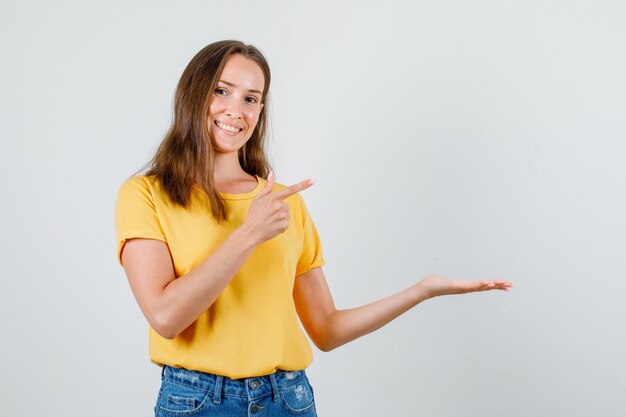 Giovane donna che punta a lato con il palmo aperto in t-shirt, pantaloncini e sembra felice. vista frontale.