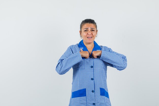 Giovane donna che punta a se stessa in camicia del pigiama a quadretti blu e sembra carina, vista frontale.