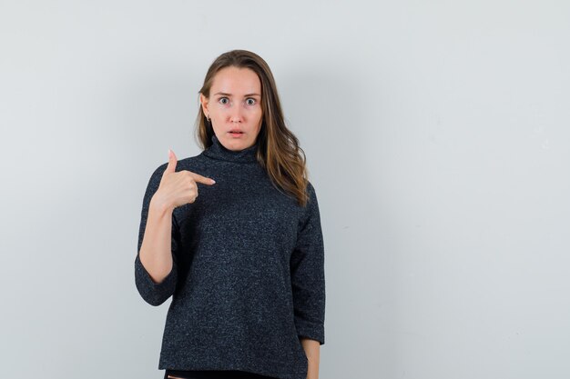 Giovane donna che punta a se stessa in camicia e che sembra perplessa. vista frontale.