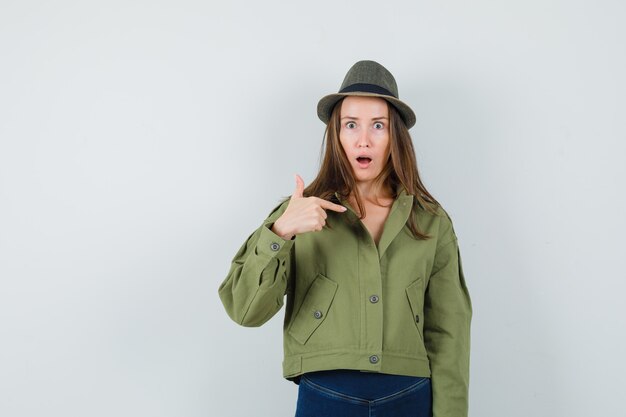 Giovane donna che punta a se stessa in giacca pantaloni cappello e guardando sorpreso