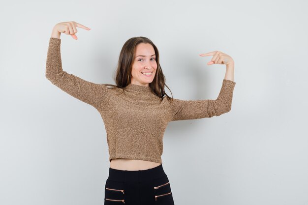 Giovane donna che punta a se stessa in maglione dorato e pantaloni neri e sembra felice, vista frontale.