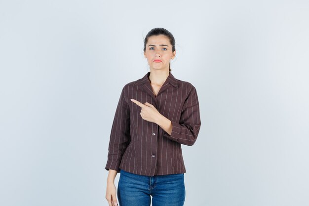 Giovane donna che punta a sinistra con il dito indice, incurvando le labbra in camicia a righe, jeans e sembra sconvolta. vista frontale.