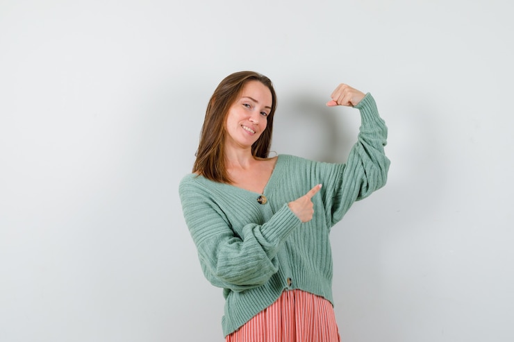 Foto gratuito giovane donna che punta ai muscoli del braccio in cardigan, gonna e che sembra allegra. vista frontale.