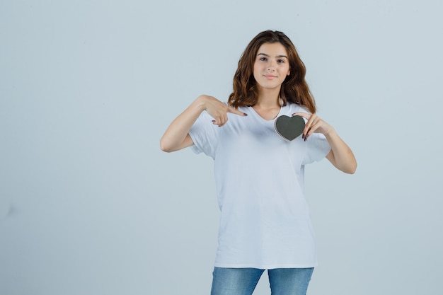 Giovane donna che punta al contenitore di regalo in maglietta bianca, jeans e che sembra felice. vista frontale.