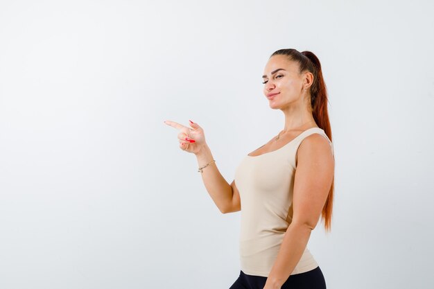 Giovane donna che punta al lato sinistro in canottiera beige e sembra fiduciosa, vista frontale.