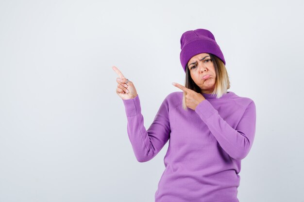 Giovane donna che punta all'angolo in alto a sinistra in maglione viola, berretto e sembra delusa, vista frontale.