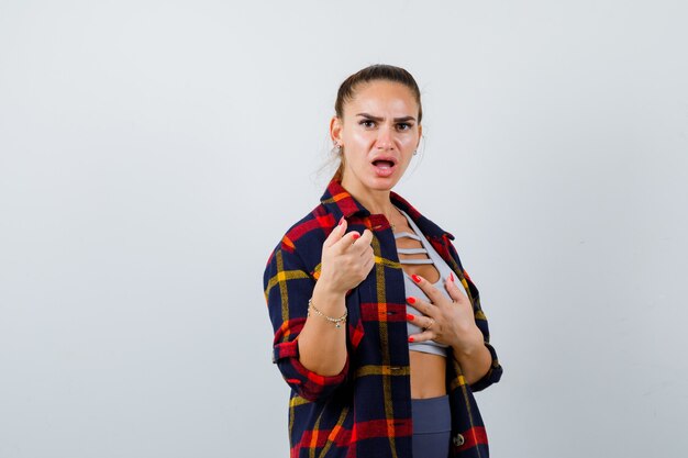 Giovane donna che punta alla telecamera in crop top, camicia a scacchi, pantaloni e sguardo perplesso, vista frontale.