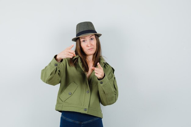 Giovane donna che punta alla telecamera in giacca pantaloni cappello e guardando esitante
