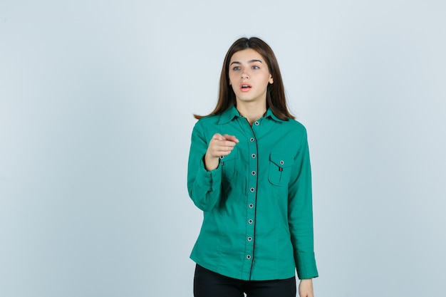 Giovane donna che punta alla telecamera mentre guarda lontano in maglietta verde e guardando scioccato, vista frontale.