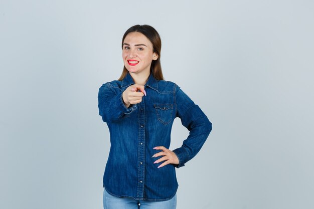 Giovane donna che punta davanti mantenendo la mano sul fianco in camicia di jeans e jeans e sembra bellissima