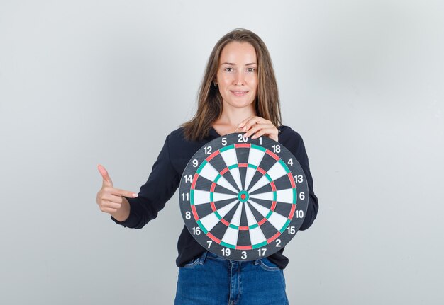 Giovane donna che punta il dito al bersaglio in camicia nera, pantaloncini di jeans e sembra allegro