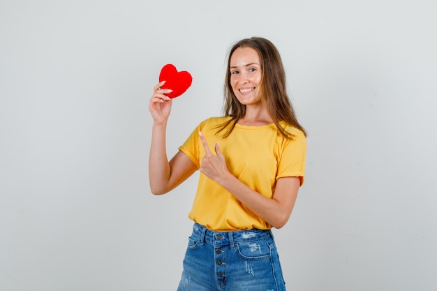 Giovane donna che punta il dito al cuore rosso in maglietta, pantaloncini e sembra allegro