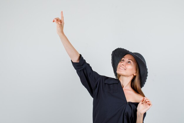 Giovane donna che punta il dito verso l'alto in camicia, cappello e sguardo allegro