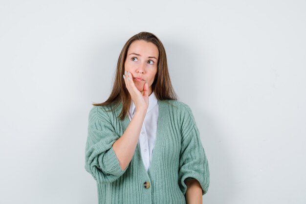 Giovane donna che puntella il mento a portata di mano in camicia, cardigan e guarda con attenzione. vista frontale.