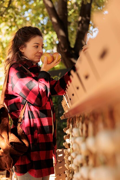 Giovane donna che sente l'odore delle mele mentre fa shopping al mercato degli agricoltori locali, acquirente femminile in piedi vicino alla bancarella che sceglie frutta e verdura fresca biologica coltivata localmente durante la visita al festival della fiera del raccolto