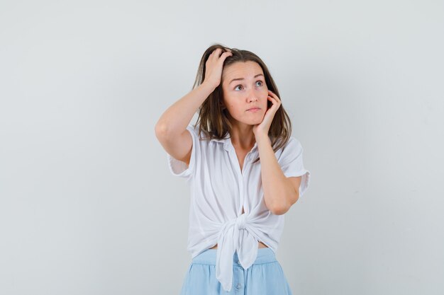 Giovane donna che si appoggia la guancia sul palmo mentre si gratta la testa in camicetta bianca e gonna azzurra e sembra allegra