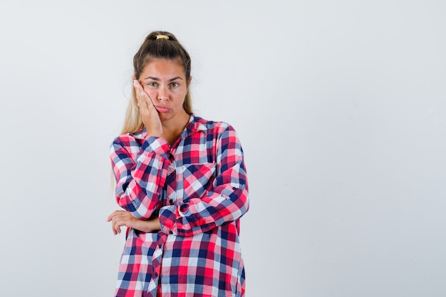 Giovane donna che si appoggia sulla guancia sul palmo della mano in camicia a quadri e guardando sconvolto, vista frontale.