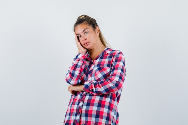 Giovane donna che si appoggia sulla guancia sul palmo della mano in camicia a quadri e sembra ragionevole. vista frontale.