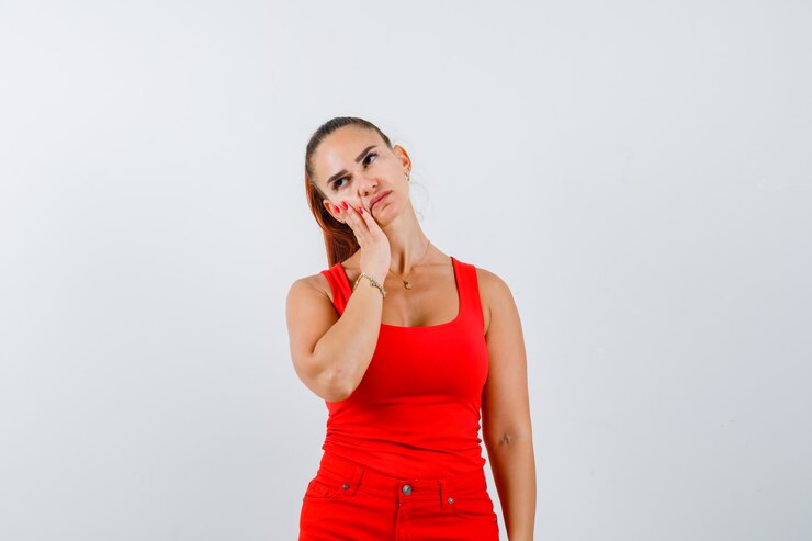 Foto gratuito giovane donna che si appoggia sulla guancia sul palmo in canottiera rossa, pantaloni e sguardo pensieroso, vista frontale.
