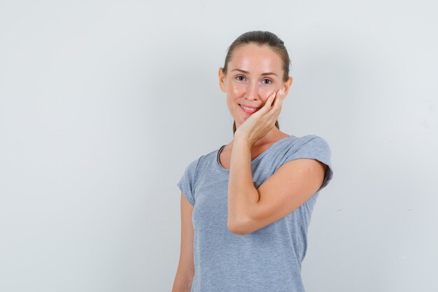 Giovane donna che si appoggia sulla guancia sul palmo sollevato in maglietta grigia e sembra felice, vista frontale.