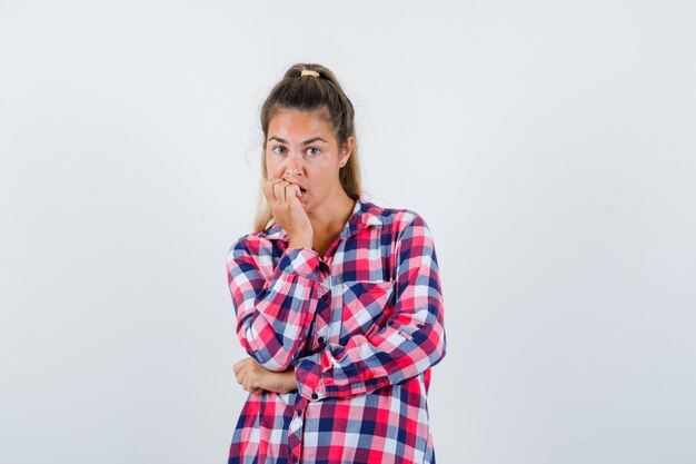 Giovane donna che si morde le unghie in camicia a quadri e sembra turbata, vista frontale.