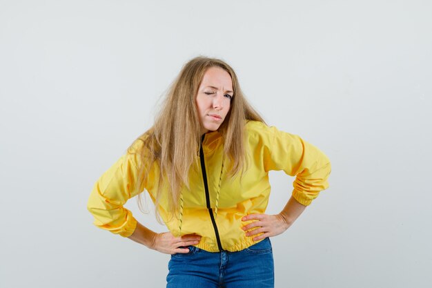 Giovane donna che si piega tenendo le mani sulla vita e ammiccante in bomber giallo e jeans blu e sembra affascinante. vista frontale.