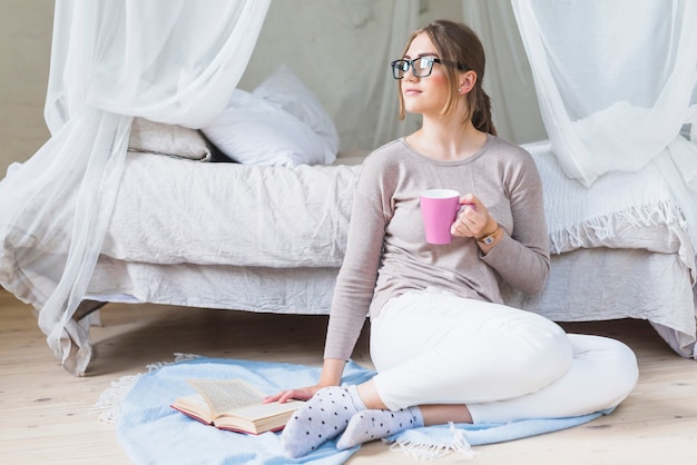 Giovane donna che si siede nella camera da letto che tiene tazza di caffè con il libro