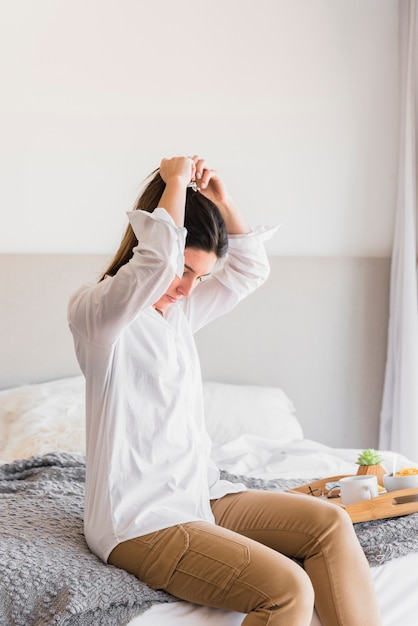 Giovane donna che si siede sul letto che lega i suoi capelli