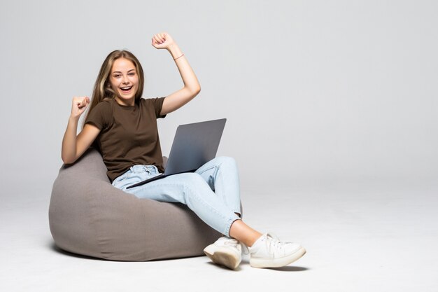 Giovane donna che si siede sul pufff con il computer portatile con il gesto di vittoria isolato sul muro bianco
