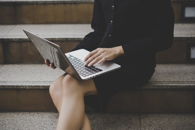 Giovane donna che si siede sulla scala per mezzo del computer portatile. Femmina che lavora al computer portatile in un esterno