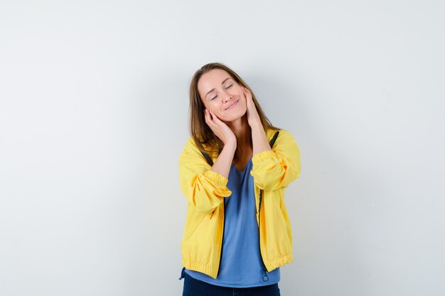 Giovane donna che si tiene per mano sulle guance, chiudendo gli occhi in maglietta, giacca e guardando serena. vista frontale.