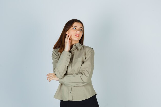 Giovane donna che si tocca la guancia mentre distoglie lo sguardo in camicia, gonna e sembra pensierosa, vista frontale.