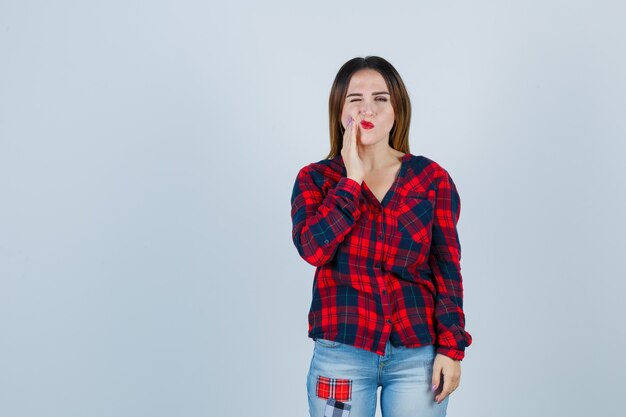 Giovane donna che soffre di mal di denti in camicia a quadri, jeans e sembra indisposta, vista frontale.