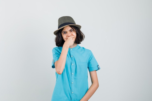 Giovane donna che soffre di mal di denti in camicia blu, cappello e sguardo turbato.