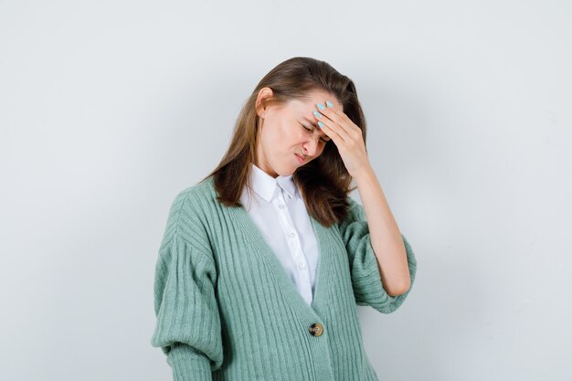 Giovane donna che soffre di mal di testa in camicia, cardigan e dall'aspetto doloroso, vista frontale.